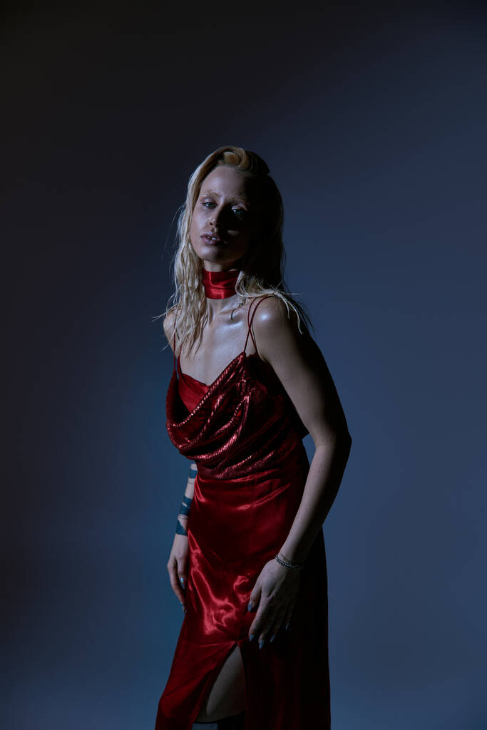 красивая женская модель с светлыми волосами в красном привлекательном платье, смотрящая на камеру на темном фоне - Фото, изображение