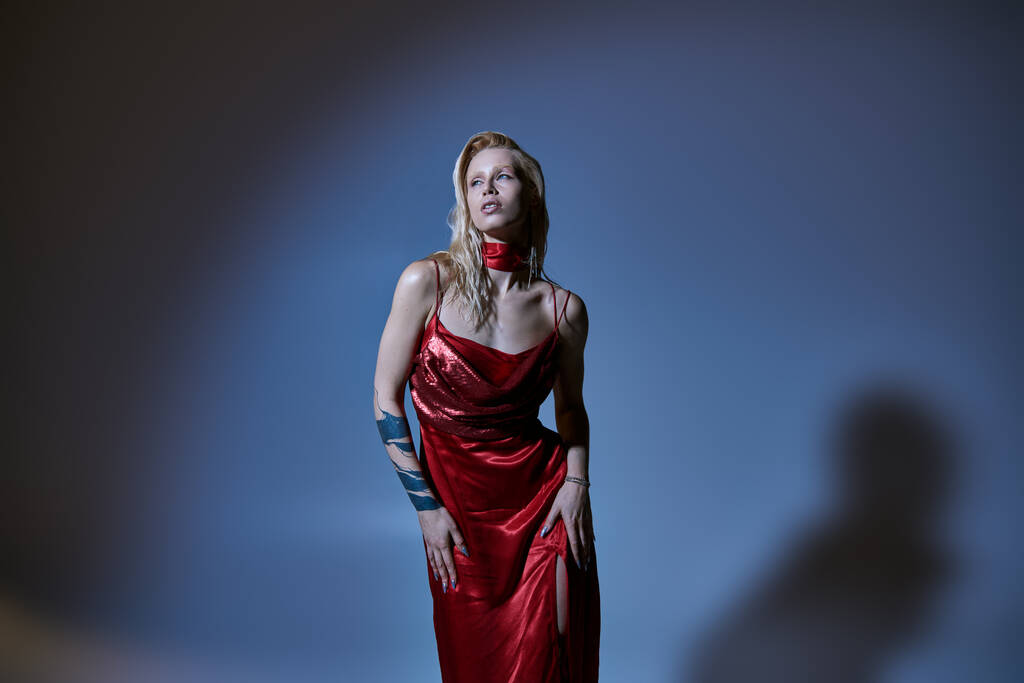 σαγηνευτική μοντέρνα γυναίκα σε κόκκινο chic φόρεμα με τατουάζ θέτοντας και κοιτάζοντας μακριά σε σκούρο φόντο - Φωτογραφία, εικόνα