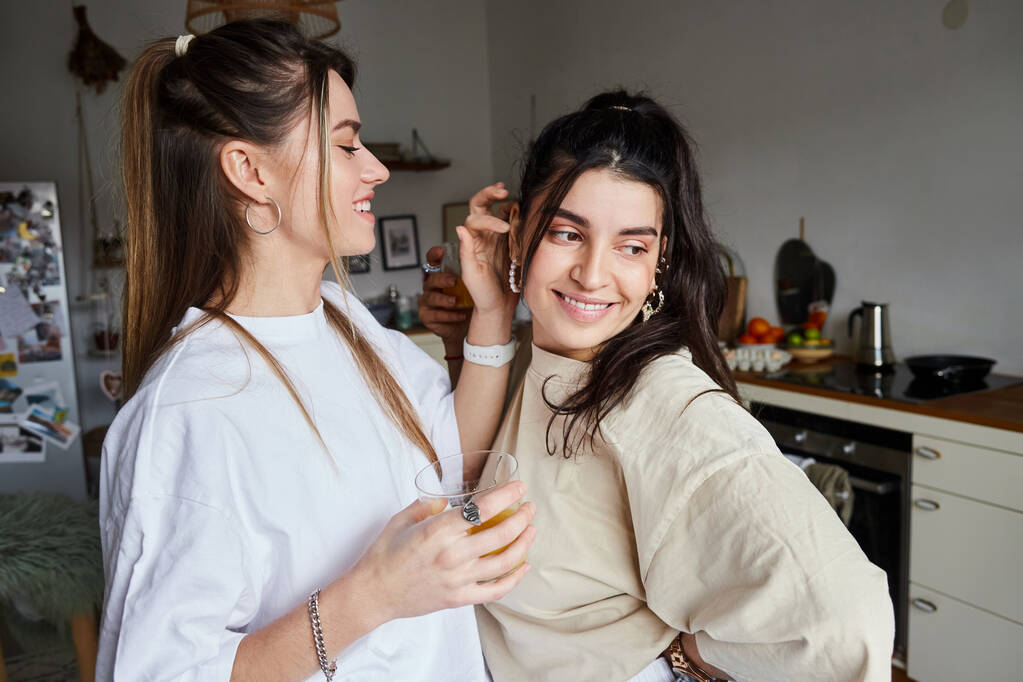 счастливая молодая женщина держит стакан свежего апельсинового сока и флирт с подругой на кухне, lgbt - Фото, изображение