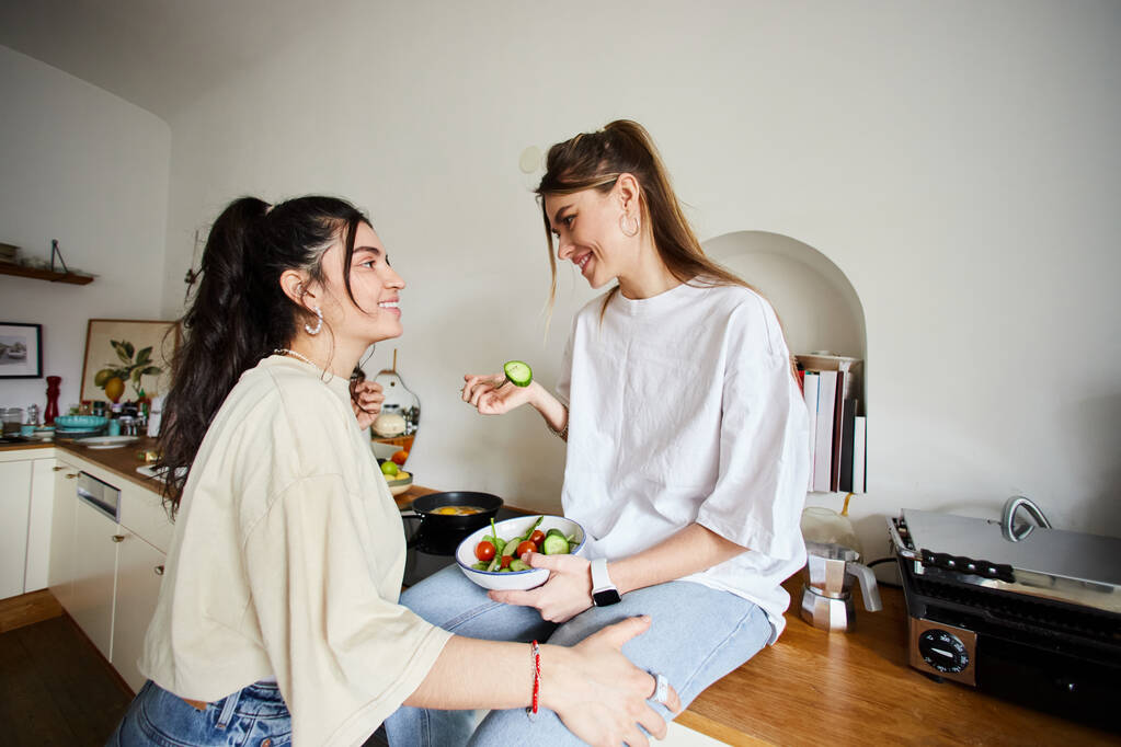 happy young lesbian woman in 20s eating salad and looking at her girlfriend in kitchen, lgbt love - Photo, Image