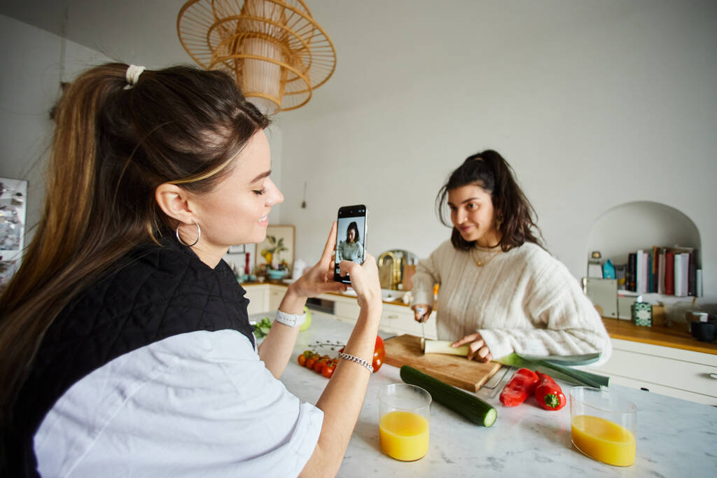 счастливая молодая лесбиянка фотографирует на смартфоне свою подружку, готовящую салат на кухне - Фото, изображение