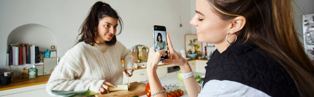 feliz joven lesbiana tomando fotos en el teléfono inteligente de su novia haciendo ensalada, pancarta - Foto, imagen