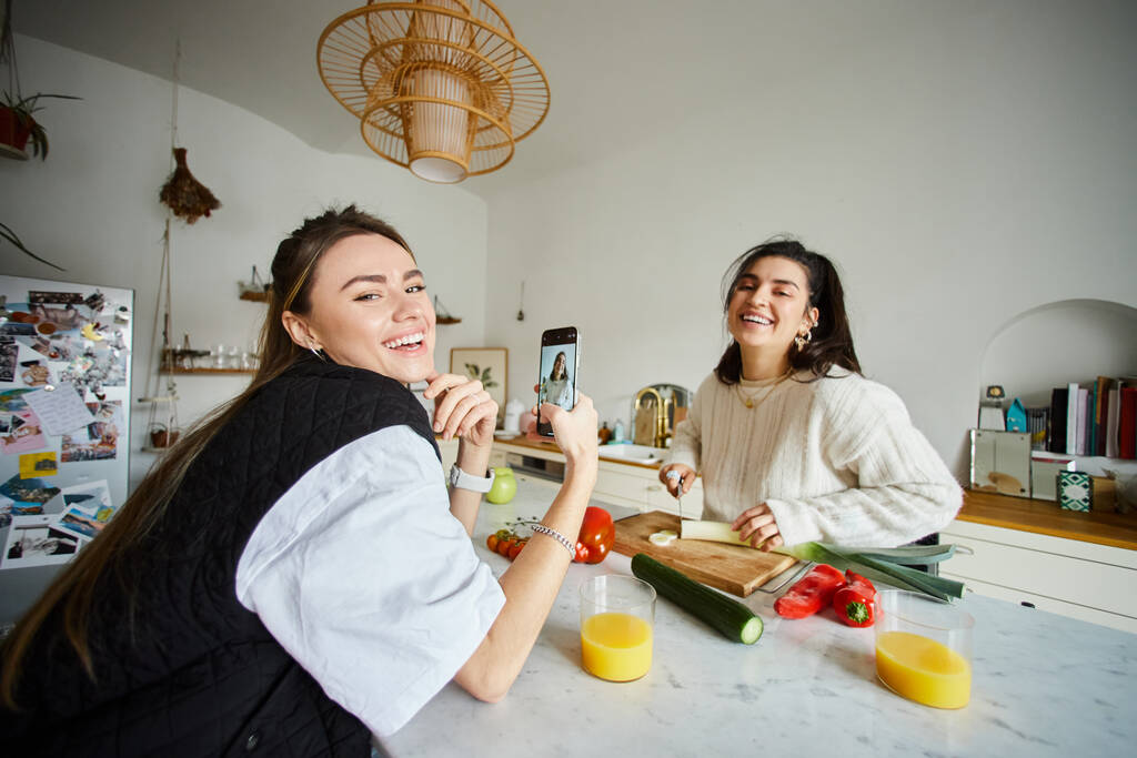 boldog fiatal leszbikus nő a 20-as években fotózza a barátnőjét, miközben salátát főz a konyhában - Fotó, kép