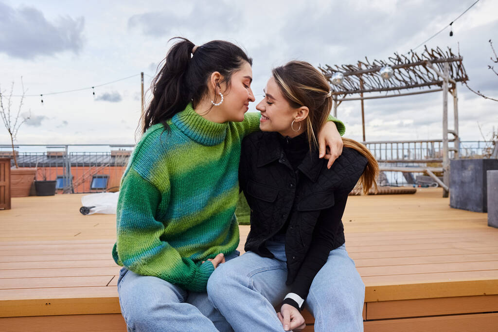 Něžná chvíle mezi veselými lesbickými ženami sedícími spolu na střeše, na pozadí města - Fotografie, Obrázek