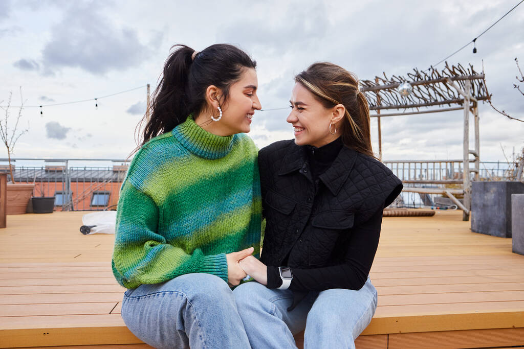 Něžná chvíle mezi pozitivními lesbickými ženami sedícími společně na střeše, na pozadí města - Fotografie, Obrázek