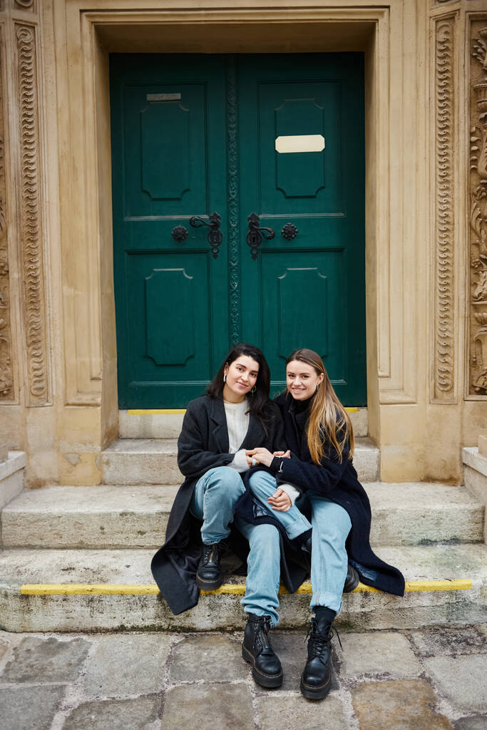 happy lesbian couple in coats sitting on stairs near historical door and holding hands of each other - Photo, Image