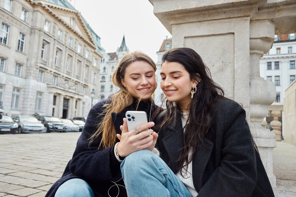 szczęśliwa i młoda lesbijka para za pomocą smartfona podczas siedzenia razem na świeżym powietrzu w Wiedeń, Austria - Zdjęcie, obraz