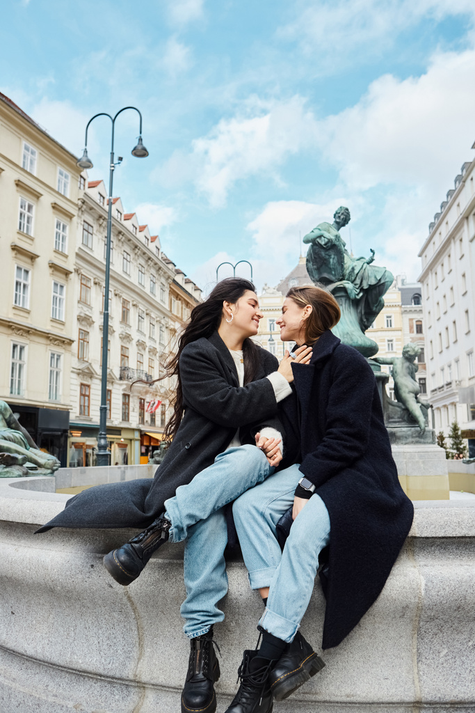 Loving lesbian couple holding hands and sitting by a fountain statue with cityscape in Vienna - Photo, Image