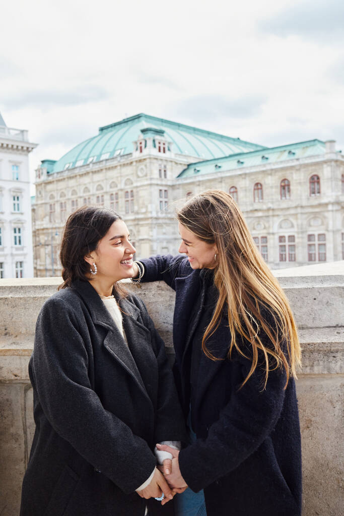 glückliches lesbisches Paar in Oberbekleidung lächelt, während es in Wien in der Nähe eines Gebäudes steht - Foto, Bild