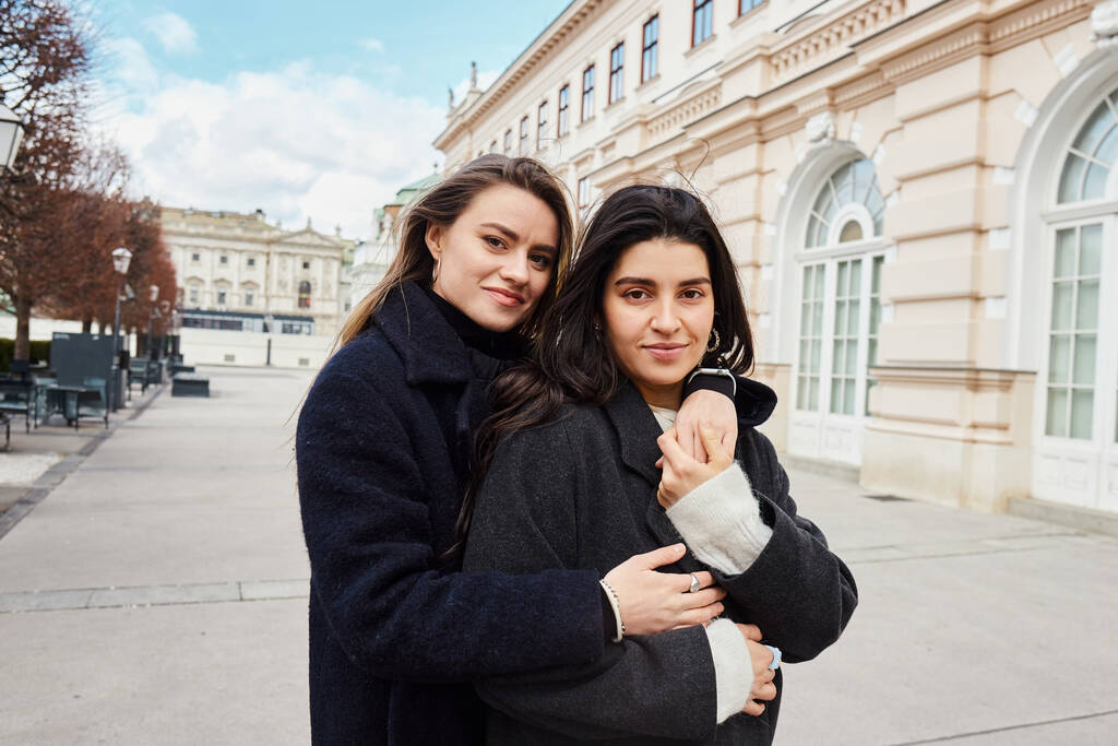 alegre lésbicas casal em outerwear sorrindo enquanto abraçando uns aos outros perto de edifício em Viena - Foto, Imagem