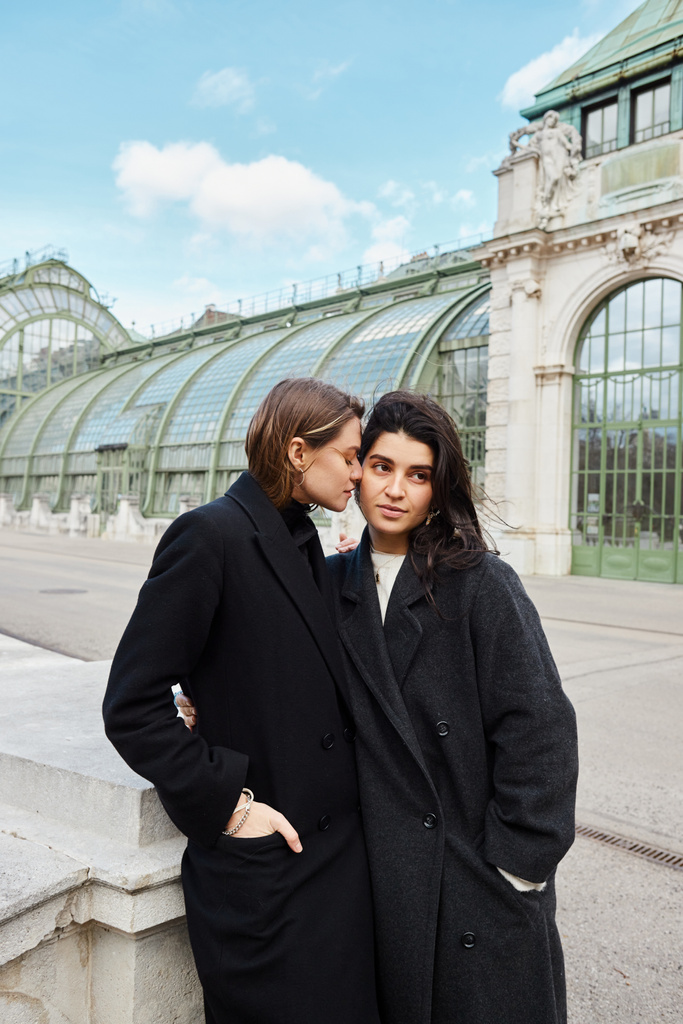 Pareja joven lesbiana en abrigos compartiendo un abrazo cercano con Palmenhaus en Viena en el fondo - Foto, Imagen