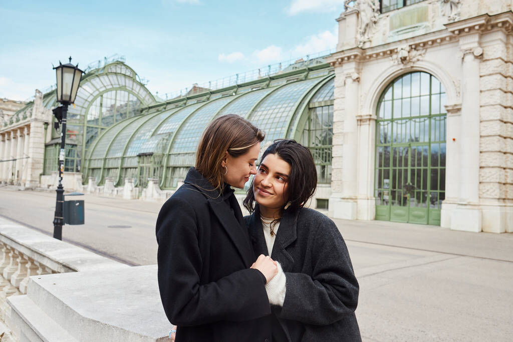 vidám leszbikus pár kabátban egymás kezét fogva a bécsi Palmenhaus közelében a háttérben - Fotó, kép