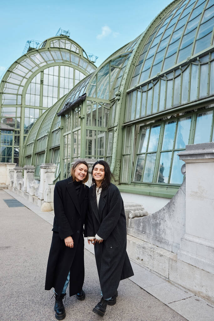 happy lesbian couple in coats holding hands while standing near Palmenhaus in Vienna on background - Photo, Image