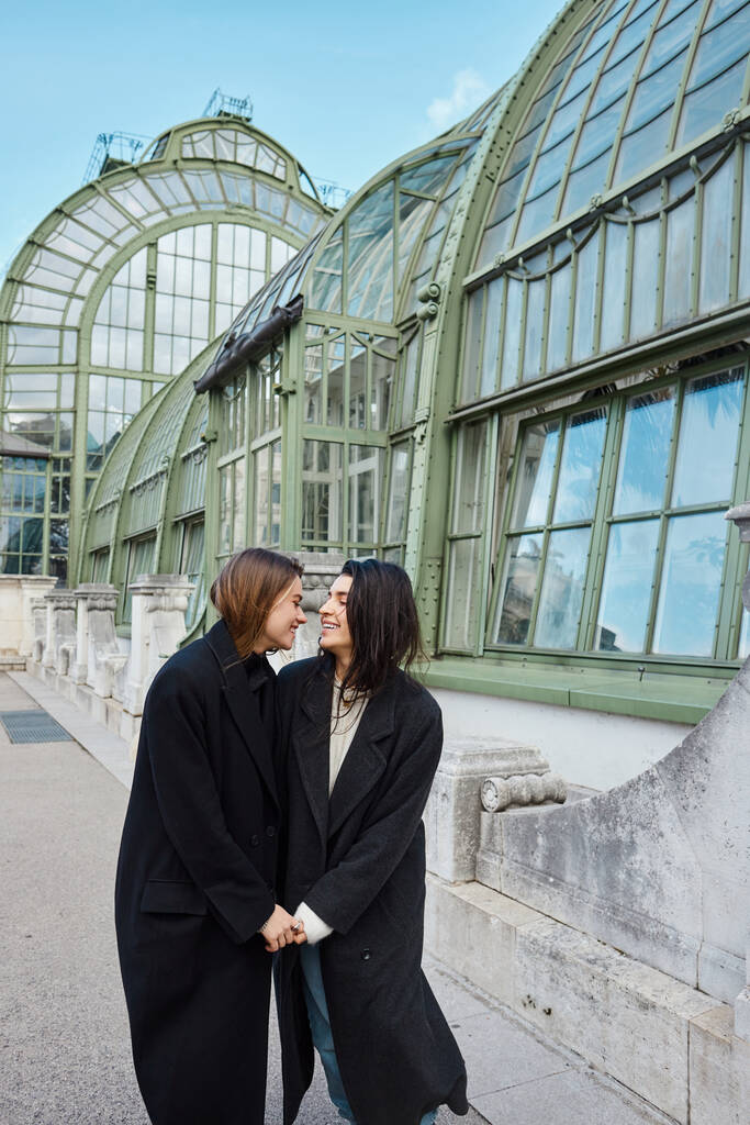lieta coppia lesbica in cappotti tenendosi per mano mentre in piedi vicino Palmenhaus a Vienna sullo sfondo - Foto, immagini