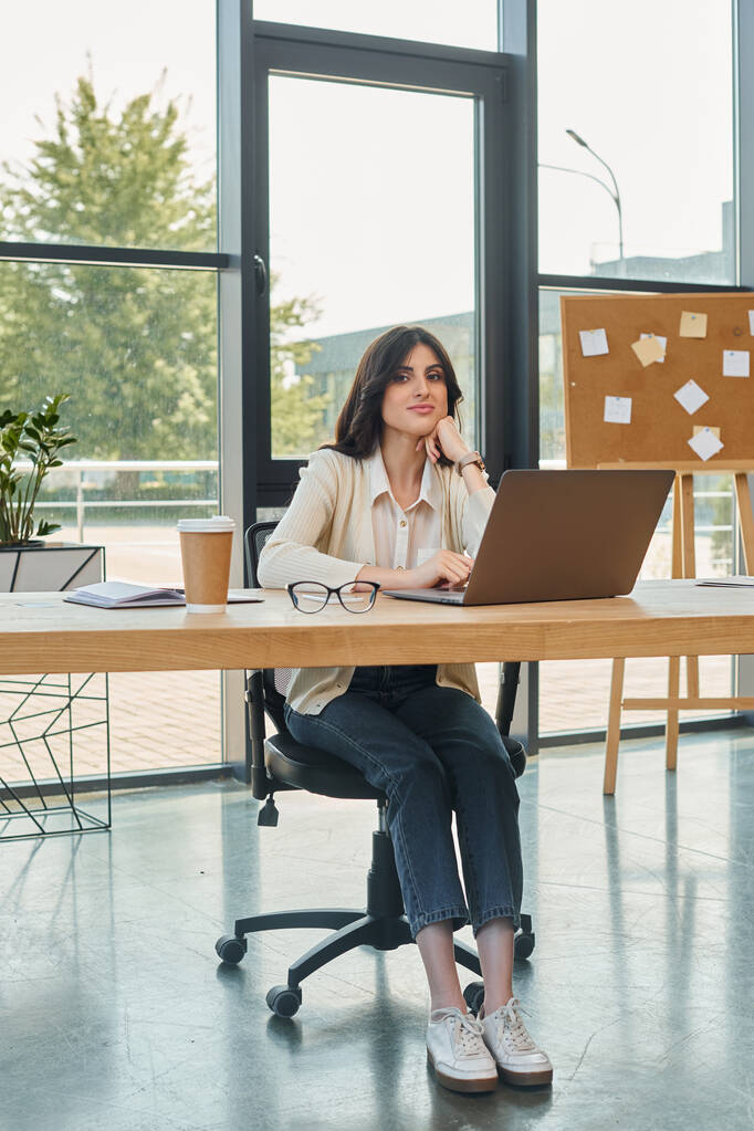 Nowoczesna bizneswoman siedzi przy biurku, skupiona na laptopie we współczesnym biurze. - Zdjęcie, obraz