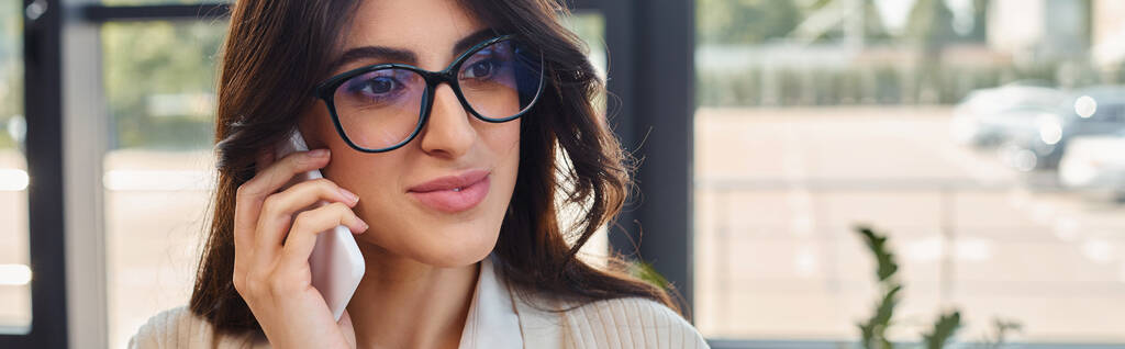 Uma empresária profissional usando óculos realiza um telefonema em um ambiente de escritório moderno perto de seu espaço de trabalho. - Foto, Imagem