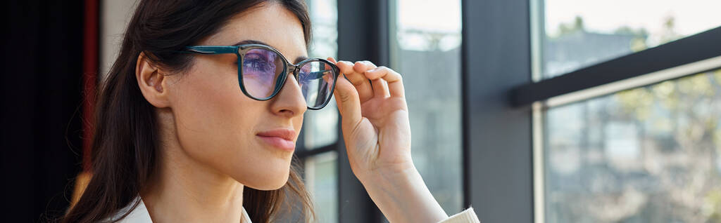 Eine selbstbewusste Geschäftsfrau mit stylischer Sonnenbrille blickt aus einem Fenster in einem modernen Büroraum und reflektiert ihren Erfolg und ihre unternehmerische Reise. - Foto, Bild