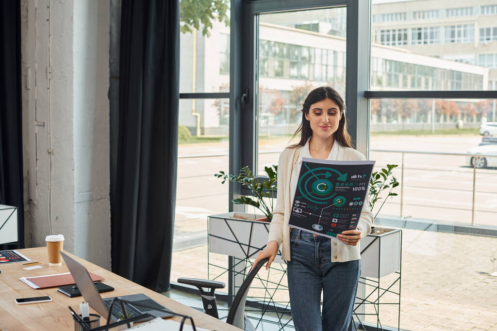 Una mujer de negocios está de pie con confianza delante de una ventana, sosteniendo gráficos con una expresión reflexiva. - Foto, imagen