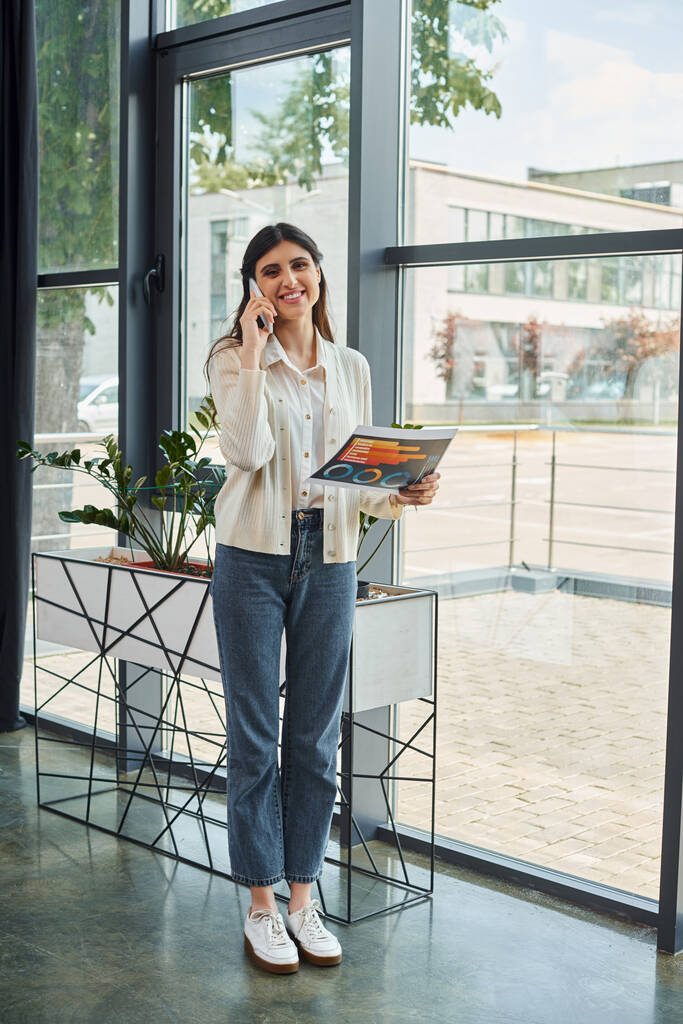 Bizneswoman stojąca przy oknie w nowoczesnym biurze, wykonująca telefon, prezentująca koncepcję franczyzy. - Zdjęcie, obraz
