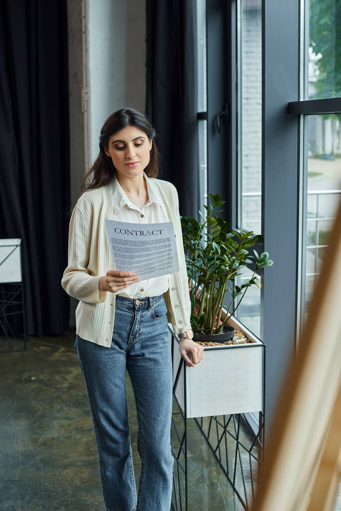 Uma empresária moderna lê um contrato em pé junto a uma janela em seu escritório, cercada por um espaço de trabalho de franquia. - Foto, Imagem