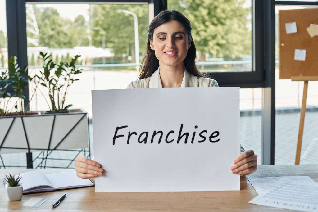Uma mulher de negócios em um escritório moderno sentado a uma mesa e segurando um sinal com cartas de franquia - Foto, Imagem