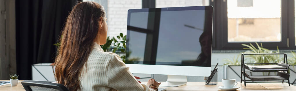 Μια επιχειρηματίας κάθεται πίσω από ένα γραφείο, εργάζεται σε έναν υπολογιστή σε ένα σύγχρονο περιβάλλον γραφείου. - Φωτογραφία, εικόνα