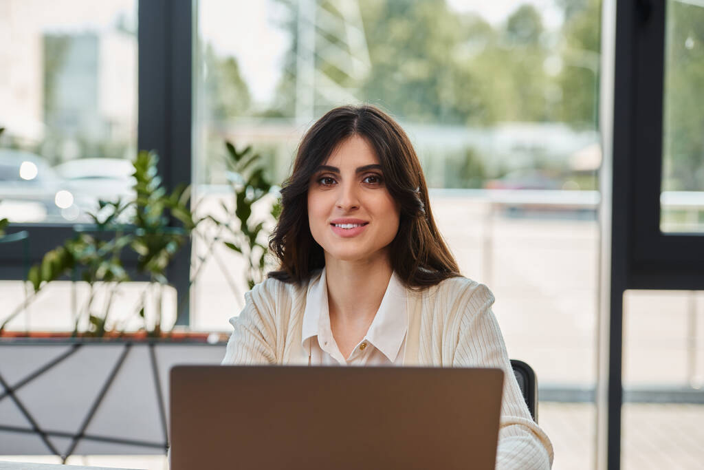 Una mujer de negocios se enfoca intensamente, sentada ante un portátil en una oficina moderna, encarnando determinación y compromiso. - Foto, imagen