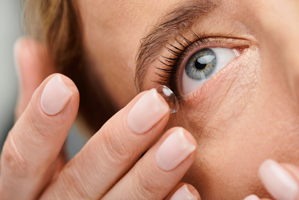 Nahaufnahme einer gut aussehenden Frau, die vorsichtig ihre Kontaktlinse trägt, während sie auf grauem Hintergrund posiert - Foto, Bild