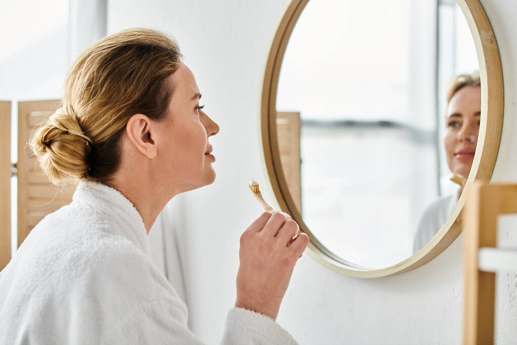 mujer rubia guapa en albornoz cepillándose los dientes delante del espejo en su baño - Foto, imagen