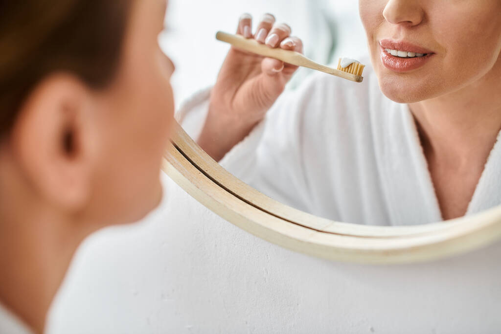 vista recortada de mujer adulta en albornoz blanco poniendo pasta de dientes en su cepillo cerca del espejo - Foto, imagen