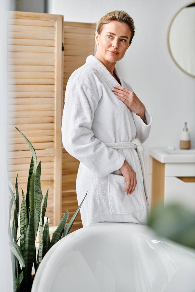 boa aparência mulher feliz com cabelo loiro em roupão aconchegante branco posando ao lado de sua banheira - Foto, Imagem