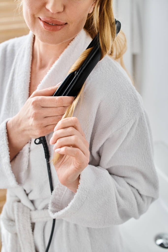 обрезанный вид на радостную блондинку в удобном халате с плоским утюгом на волосах в ванной комнате - Фото, изображение