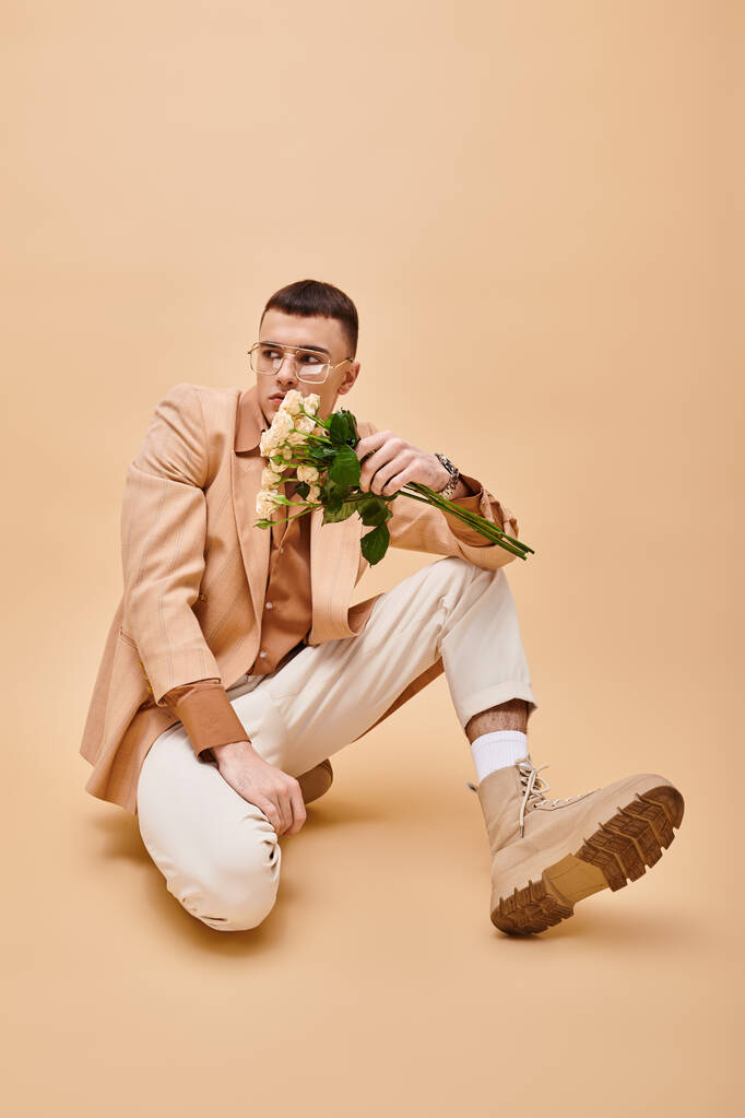 Fashion shot van de mens in beige jas en bril zitten met bloemen op perziken beige achtergrond - Foto, afbeelding