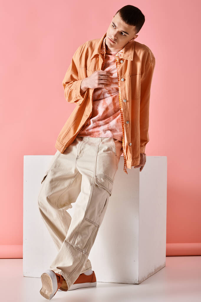Fashion image of stylish man in beige shirt, pants and boots on white cube on pink backdrop - Photo, Image