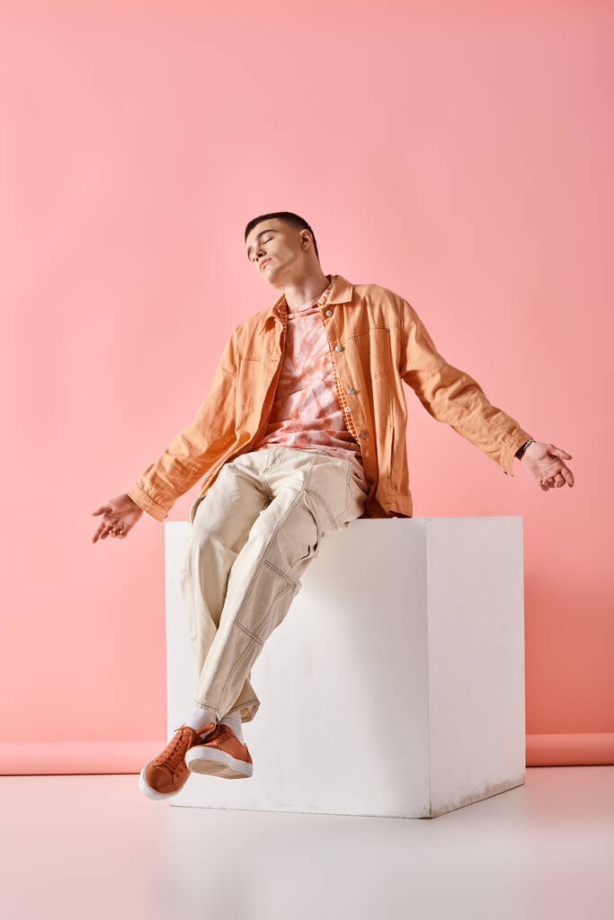 Homme élégant en tenue beige tenant les bras ouverts et assis sur un cube blanc sur fond rose - Photo, image