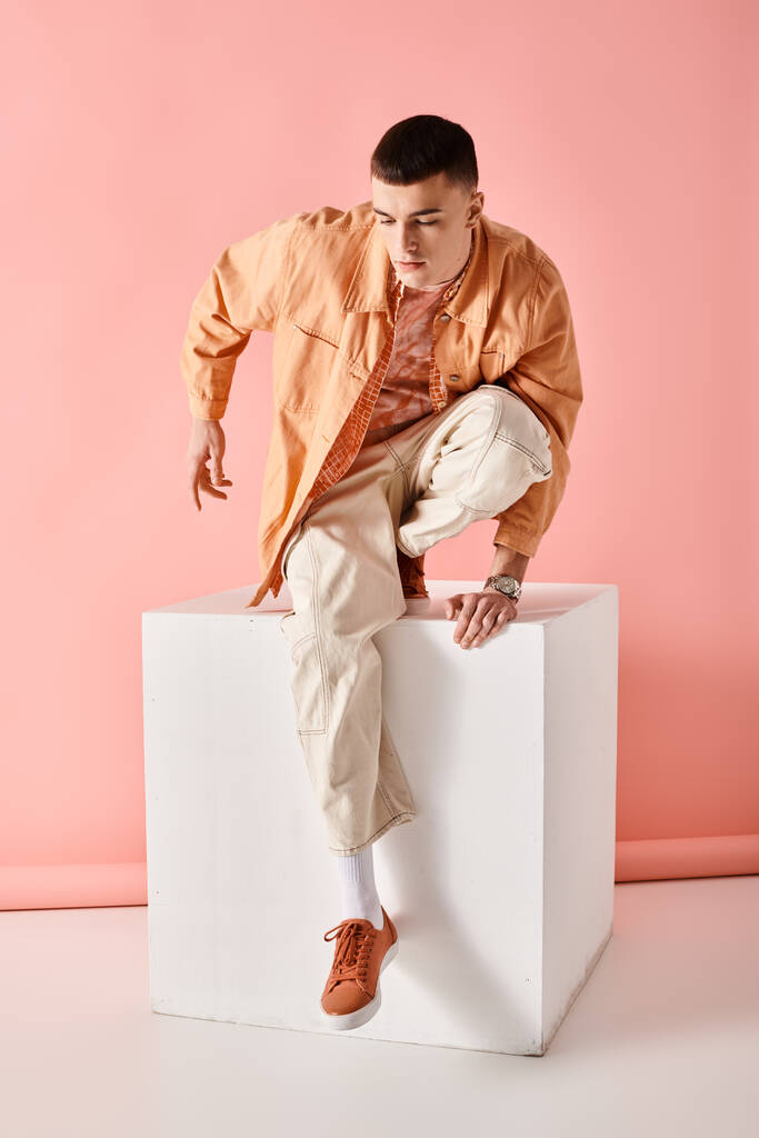 Красивый мужчина в бежевом наряде смотрит вниз и позирует на белом кубе на розовом фоне - Фото, изображение