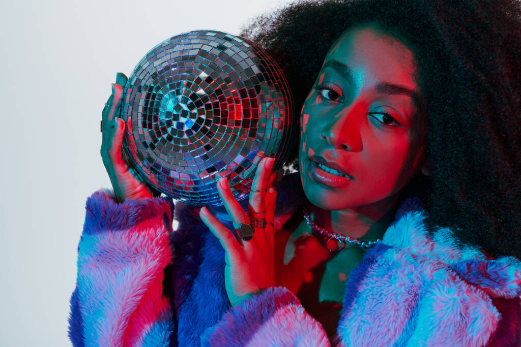 appealing stylish african american woman in faux fur posing with disco ball and looking at camera - Photo, Image