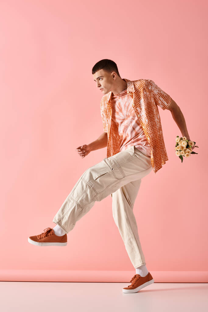 Full length image of fashionable man in layered outfit holding flowers on pink backdrop - Photo, Image