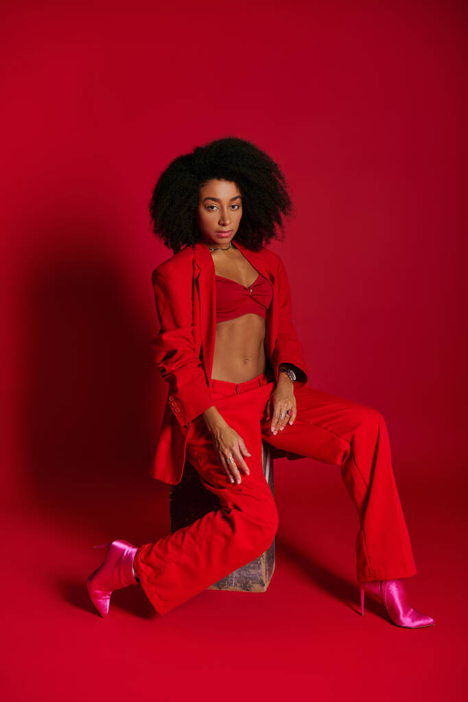 attraente donna afro-americana sexy in abito rosso seduto e guardando la fotocamera, concetto di moda - Foto, immagini