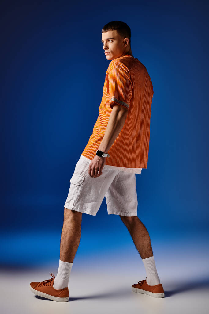 Largura completa vista lateral de hombre guapo en camisa naranja y pantalones cortos blancos posando sobre fondo azul - Foto, imagen