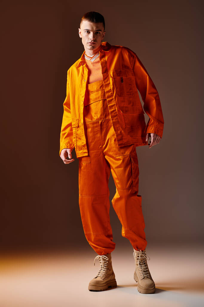 Full length portret van modieuze man in oranje jumpsuit en jas poseren op bruine achtergrond - Foto, afbeelding