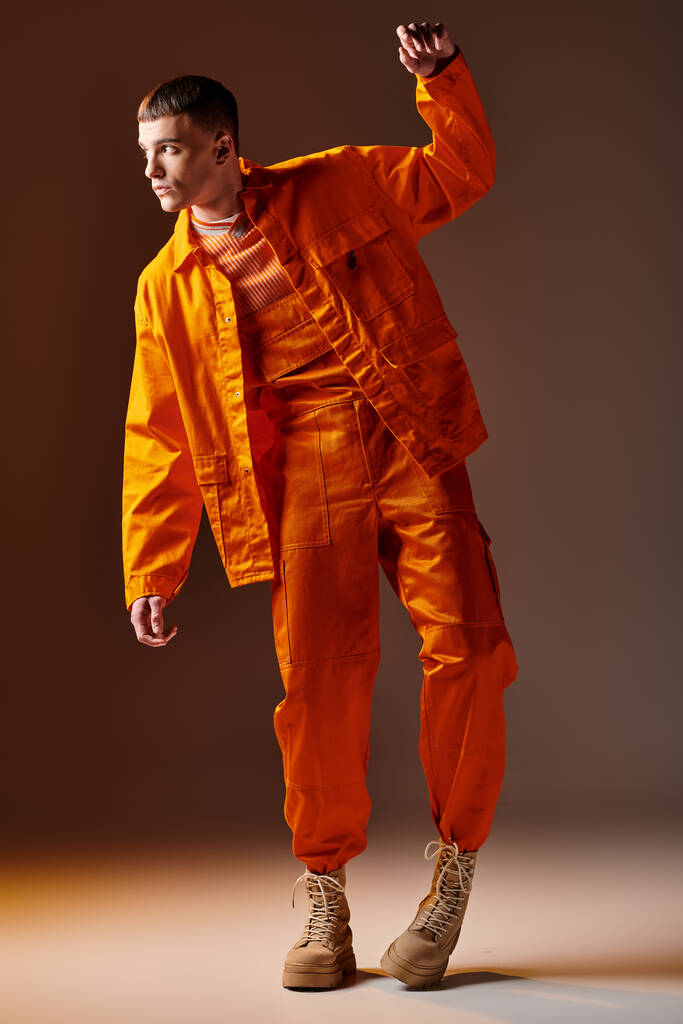 Полная длина снимка стильного мужчины в оранжевом комбинезоне и куртке, позирующего на коричневом фоне - Фото, изображение