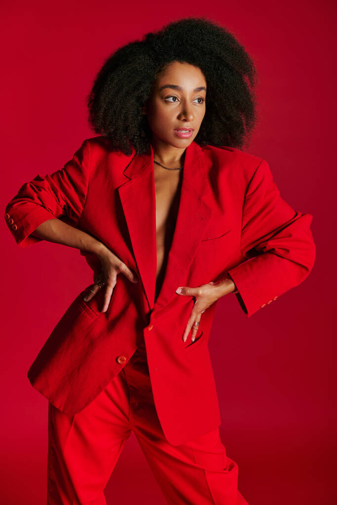 σαγηνευτικό αφρικανικό αμερικανικό θηλυκό μοντέλο σε κόκκινο σακάκι κοιτάζοντας μακριά στο κόκκινο φόντο, έννοια της μόδας - Φωτογραφία, εικόνα