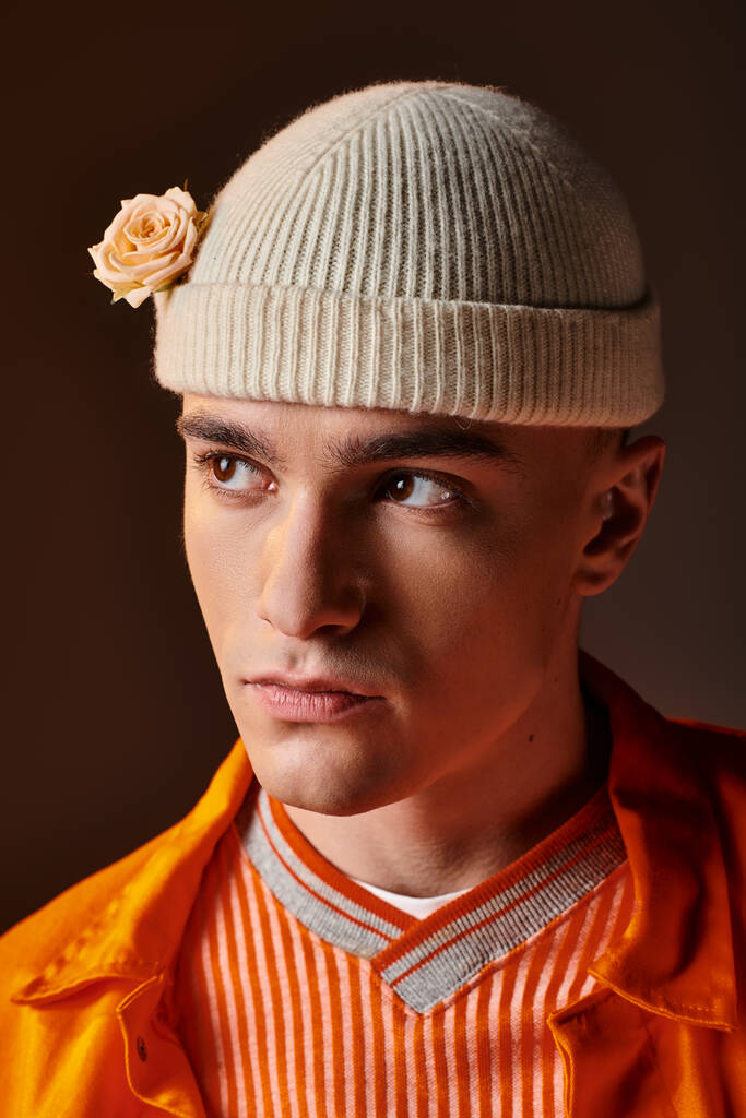 Portrait of handsome man in orange outfit wearing beige beanie with flower on brown background - Photo, Image