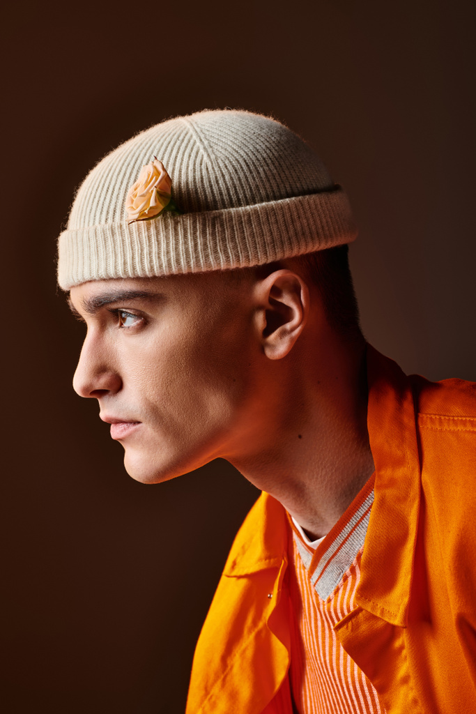 Side view of stylish man in orange outfit wearing beige beanie with flower in studio - Photo, Image