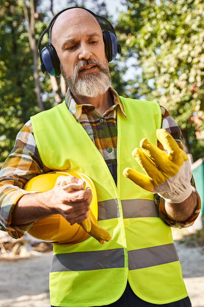 Güvenlik eldiveni ve yelek giyen, kulaklık ve kaskla poz veren çalışkan bir yazlık inşaatçısı. - Fotoğraf, Görsel