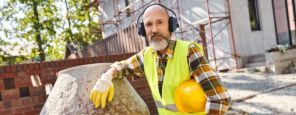 alegre constructor de cabaña en guantes de seguridad y chaleco con auriculares y casco sonriendo a la cámara, pancarta - Foto, Imagen