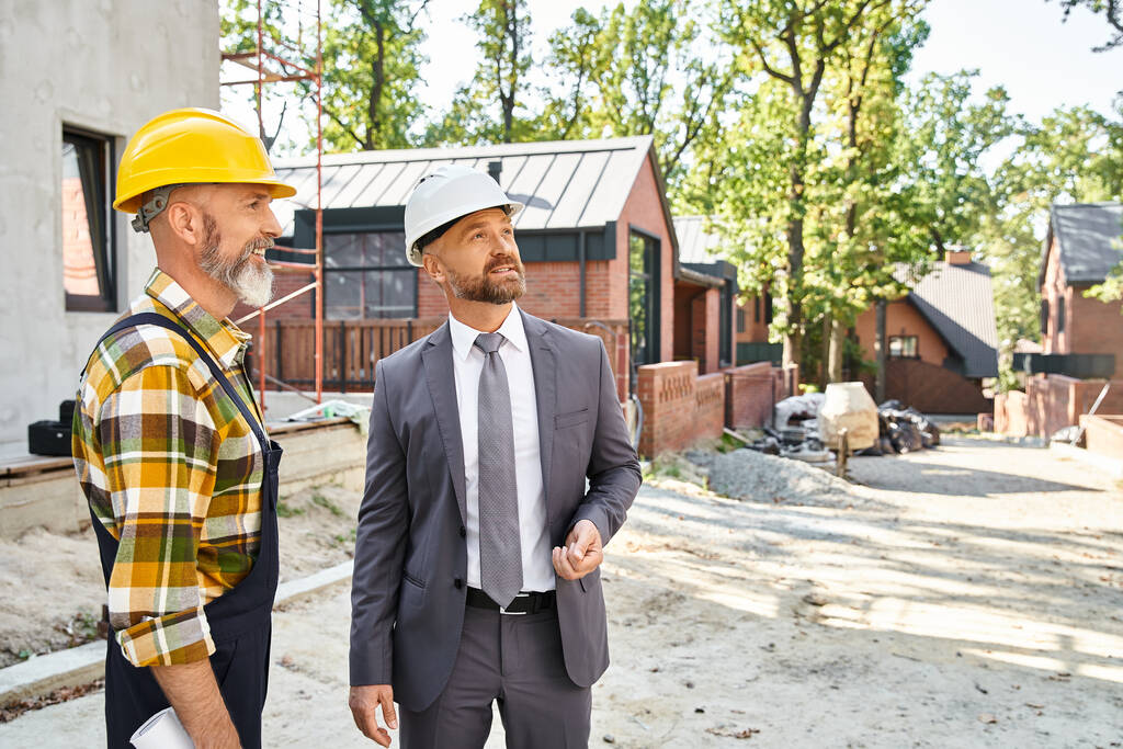 alegre constructor de cabañas y arquitecto en traje y overoles con plano discutiendo la construcción - Foto, imagen