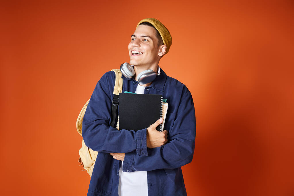 Улыбающийся молодой студент в наушниках и в жёлтой шляпе держит рюкзак и обнимает записки - Фото, изображение