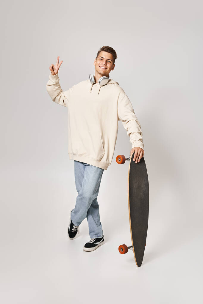 красивый студент в легкой толстовке и наушниках, стоящий со скейтбордом и показывающий знак мира - Фото, изображение